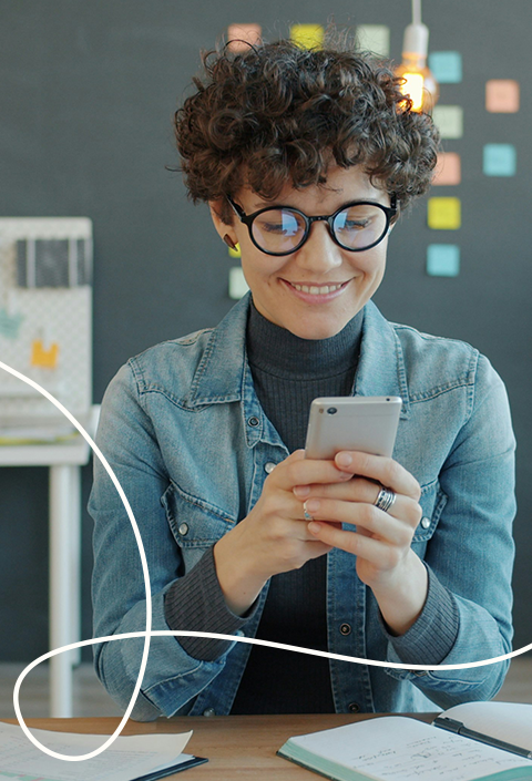 Bild einer lockigen Frau die an einem Schreibtisch sitzt und in ihr Smartphone schaut dazu eine helle geschwungene Linie in der unteren linken Ecke als Gestaltungselement | Kremer Signature Design