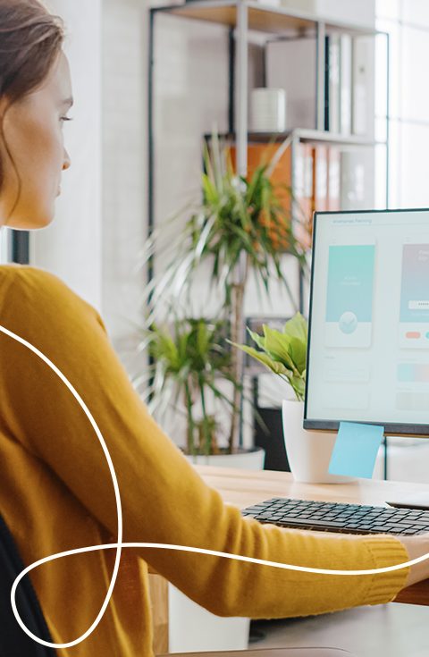 Foto einer Frau mit gelben Pullover die am Schreibtisch sitzt und am Computer arbeitet dazu in der linken Ecke eine helle geschwungene Linie als Gestaltungselement, Symbolisches Bild für Grafikdesign | Kremer Signature Design